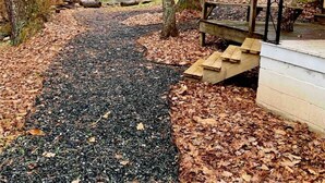 A gravel path leading to a wooden bench and a small wooden structure, surrounded by trees and a building with string lights. - Creekside Cabin skiing/hiking & Appalachian Trail. (Green Mountain)