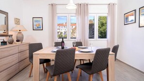 A dining area with a wooden table, six chairs, a bottle of wine, and a view of buildings through large windows. - Holiday house Sunshine in center of Jelsa (Jelsa)