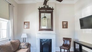 Een woonkamer met een open haard, een bank, een fauteuil, een televisie en lijstwerken aan de wanden.