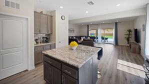 A modern kitchen with granite countertops, a central island, and a dining area with a view. - Family Paradise Saltwater Pool & Spa Game Room Fire Pit  EV Charger  Poker Table (Desert Hot Springs)