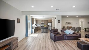 A modern living room with a flat-screen TV, leather sofa, and a dining area with a kitchen in the background. - Family Paradise Saltwater Pool & Spa Game Room Fire Pit  EV Charger  Poker Table (Desert Hot Springs)