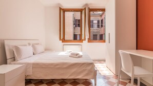 Una camera da letto con un letto, una scrivania e una sedia. Si vede una finestra con vista sugli edifici.