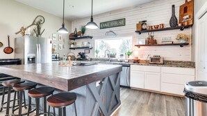 Une cuisine dotée d’un grand îlot central, de tabourets de bar, de luminaires suspendus et d’étagères en bois où sont rangés divers ustensiles de cuisine.