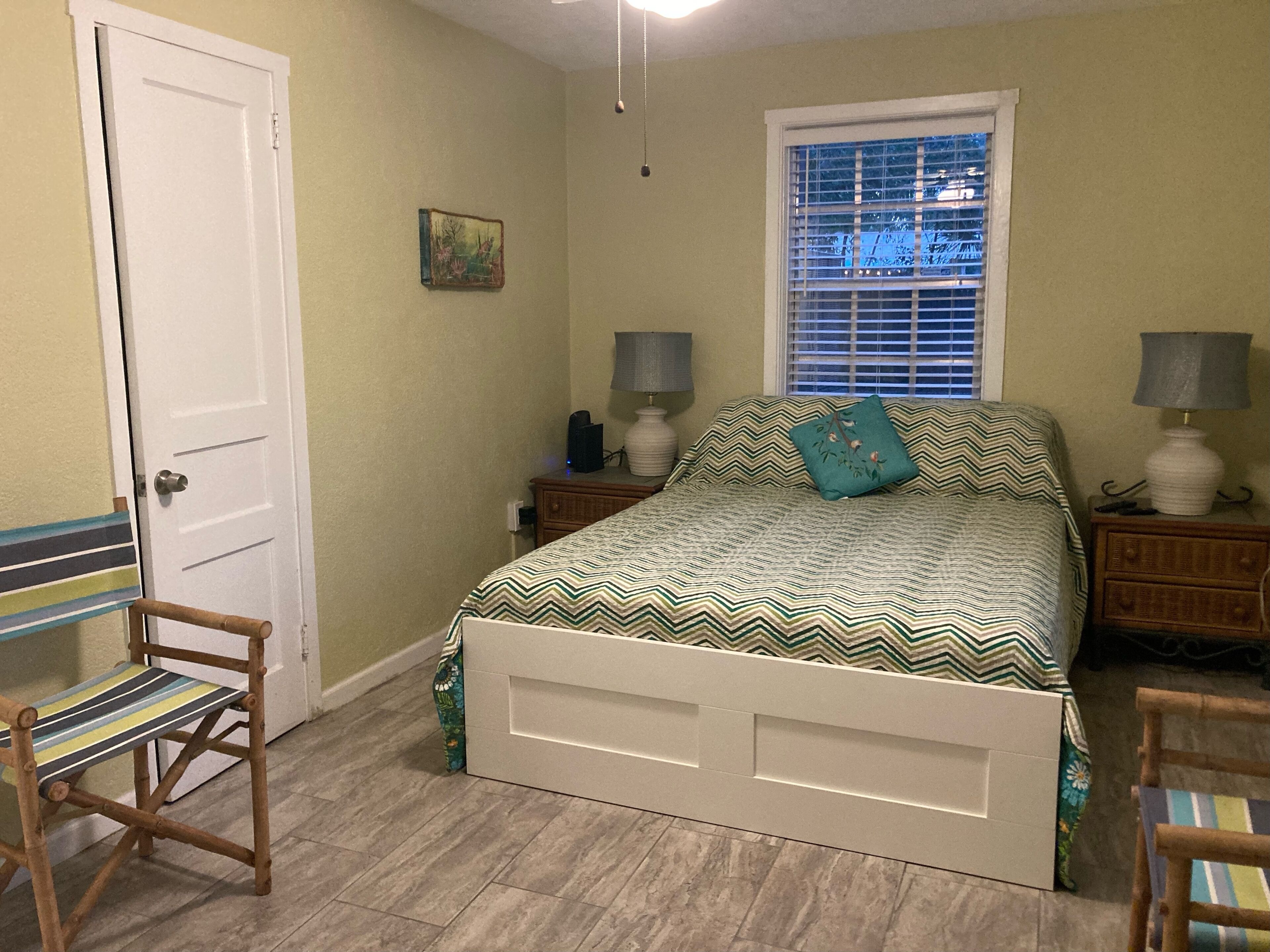 A bedroom with a bed, two nightstands, a window with blinds, a door, and a chair.
