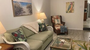 Una sala de estar con un sofá verde, una silla de mimbre, una mesa de centro de vidrio y un cuadro de un loro en la pared.