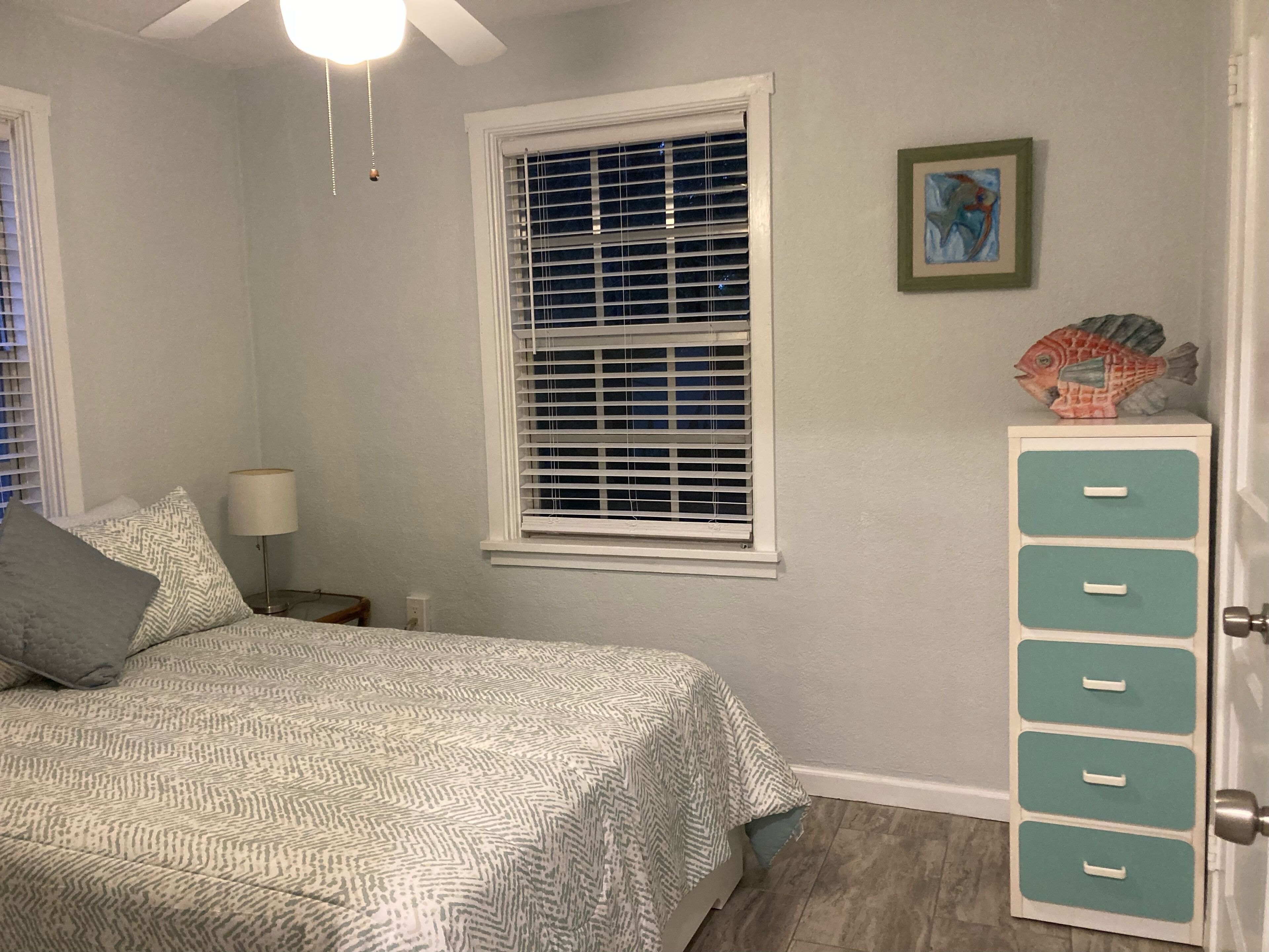 A bedroom with a bed, a nightstand, a lamp, a window with blinds, a fish decoration, and a dresser with drawers.