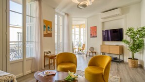 A modern living room with large windows, a flat-screen TV, a sofa, a coffee table with flowers, and a potted plant. - Sublime flat, city centre, modern, balconies, air conditioning, refurbished (Nice)