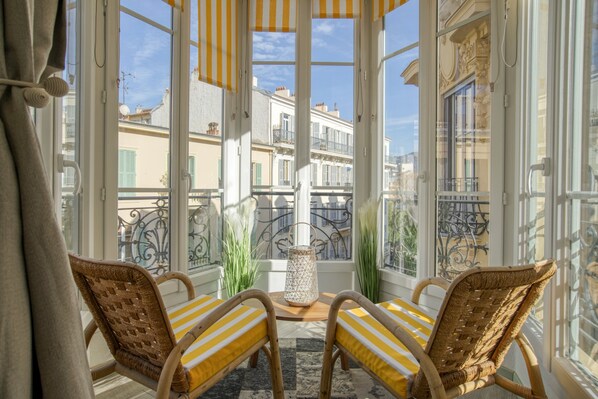 Ein Zimmer mit Korbstühlen, einem Tisch und Blick auf Gebäude durch große Fenster.