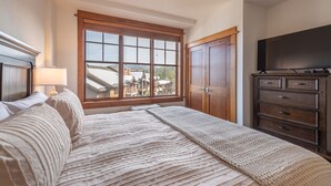 Un dormitorio con un ventanal grande, una cama con manta texturizada, un aparador de madera y un televisor en la pared.