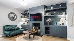 A modern living room with a green sofa, a fireplace, a TV mounted on the wall, and a bookshelf with decorative items. - Cozy 3 Bedroom Terrace Cottage in Epsom (Epsom)