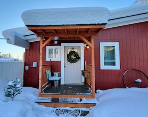 A wooden porch with a snow-covered roof, a door with a wreath, a bench, and a bicycle wheel leaning against the wall.