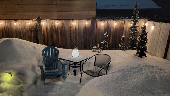 A snow-covered patio with a table and two chairs, a string of lights, and a small Christmas tree.
