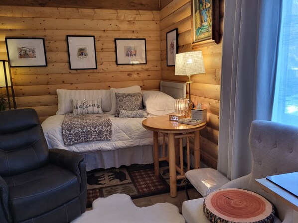 A cozy room with a bed, a round table with a lamp, a chair, and a sofa. There are framed pictures on the wall and a window with curtains.
