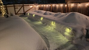 A snow-covered pathway illuminated by string lights, leading to a wooden fence.
