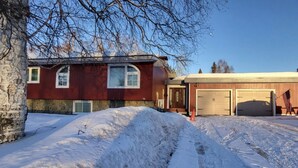 A two-story house with a stone foundation and a red exterior. There are two garage doors and a driveway with snow. - A Private City, Cabin-feel Studio for 1 Person (Fairbanks)
