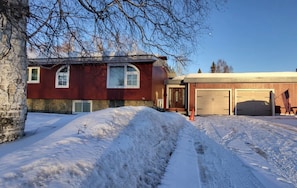A two-story house with a stone foundation and a red exterior. There are two garage doors and a driveway with snow.