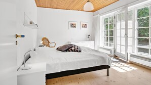 Ett sovrum med ett träpanelat tak, en säng med en svart ram, ett vitt sängbord, en flätad stol och stora fönster med utsikt över grönskan.