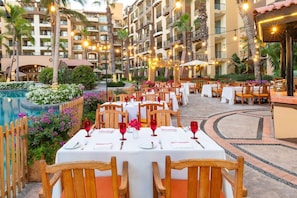 Guests can dine al fresco amidst a warm ambiance, surrounded by a pool and a multi-story building with balconies.