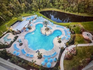 Eine Vogelperspektive zeigt einen Urlaubsort mit einem großen zentralen Pool, umgeben von Liegebereichen und einem kleinen Teich.