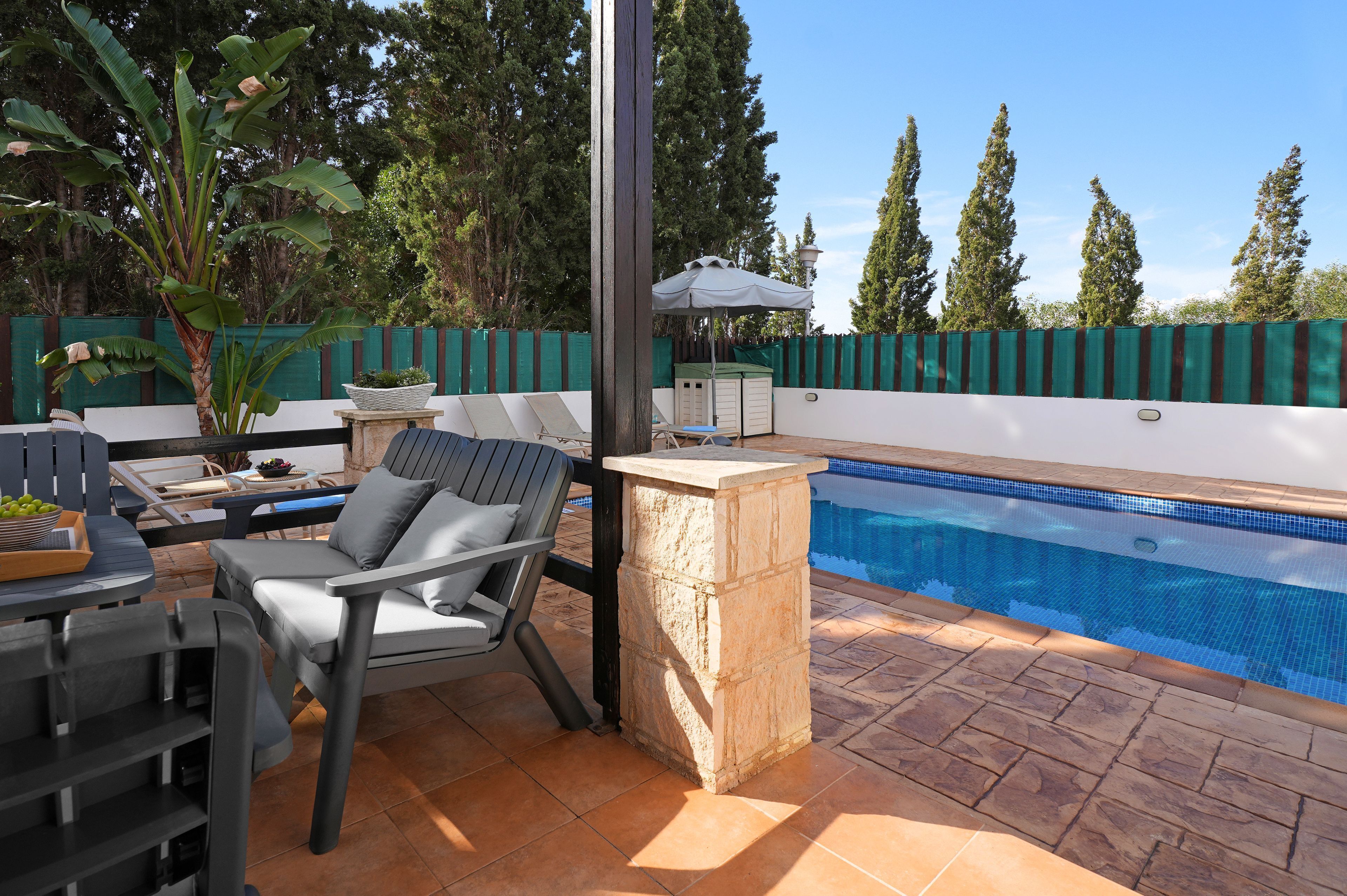 Zona junto a la piscina con un banco de piedra, sillas de descanso y una mesa pequeña con un cuenco de fruta.