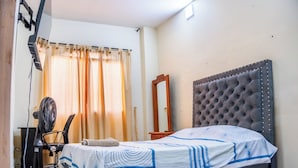 Une chambre à coucher comprenant un lit, un bureau, une chaise, un miroir et un ventilateur fixé au mur.