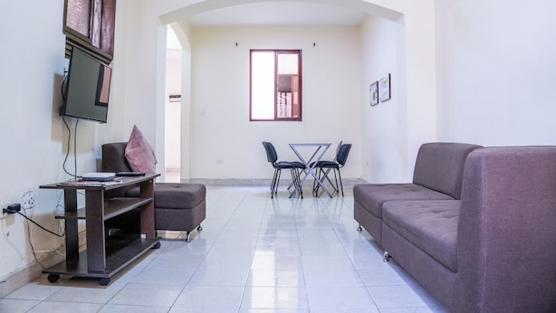 Un salon avec un canapé, un téléviseur posé sur un meuble, une table avec des chaises et une fenêtre.