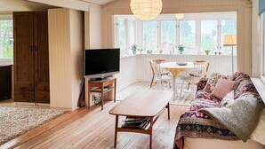 Una sala de estar acogedora con televisor, muebles de madera, una zona de comedor y ventanas amplias.