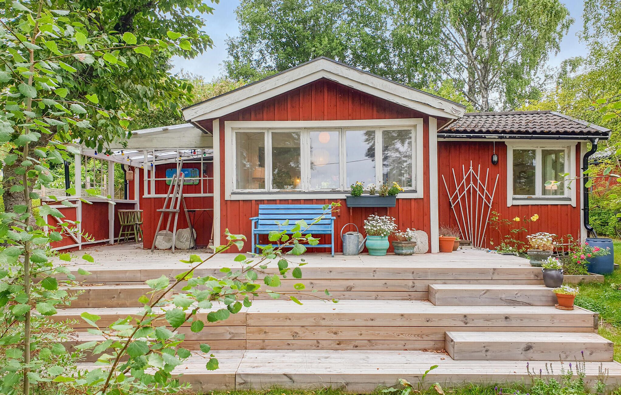 Une maison en bois rouge avec une grande fenêtre, un banc bleu et un escalier en bois menant à la porte d’entrée.