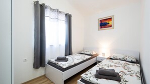 Un dormitorio pequeño y luminoso con dos camas individuales, una ventana con cortinas, una mesita de noche con lámpara y un cuadro en la pared.