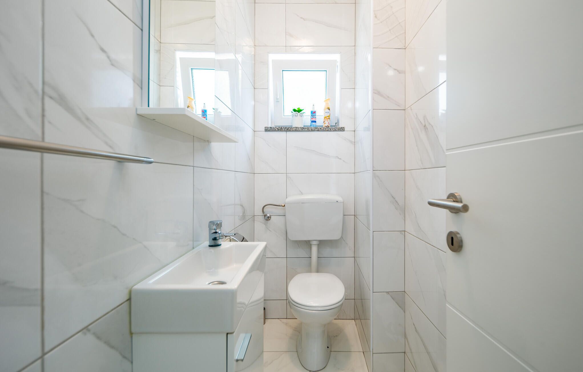 A small, modern bathroom with white marble tiles, a white toilet, a sink, and a shower area.