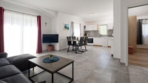 Una sala de estar moderna con un sofá, una mesa de centro y un televisor. Se observa un comedor con una mesa y sillas. La cocina se ve al fondo, con armarios blancos y un salpicadero de azulejos.