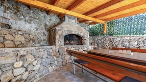 Cocina exterior de piedra con horno de pizza integrado, bancos de madera y pared de piedra.