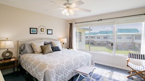 A bedroom with a large bed, a ceiling fan, a window with a view of buildings, a chair, and a nightstand with a lamp. - Island Oasis with a view (Dauphin Island)