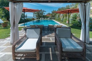 Un área junto a la piscina con sillones de mimbre, un patio de piedra y sombrillas rojas.