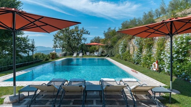 Ein Swimmingpool mit Liegestühlen und Sonnenschirmen, umgeben von viel Grün und Blick auf die Berge.