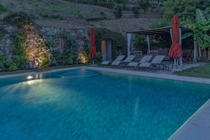Una piscina con sillas de descanso y sombrillas, un muro de piedra con plantas, y un área de estar techada.