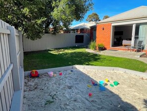 A backyard with a sandy play area, a white fence, and a brick house with a sliding glass door. - Sunbury on the Park - netflix, table tennis (Sunbury)