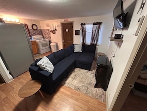 Un espacio habitable compacto con un sofá modular, una pequeña mesa redonda, una kitchenette y un televisor montado en la pared.