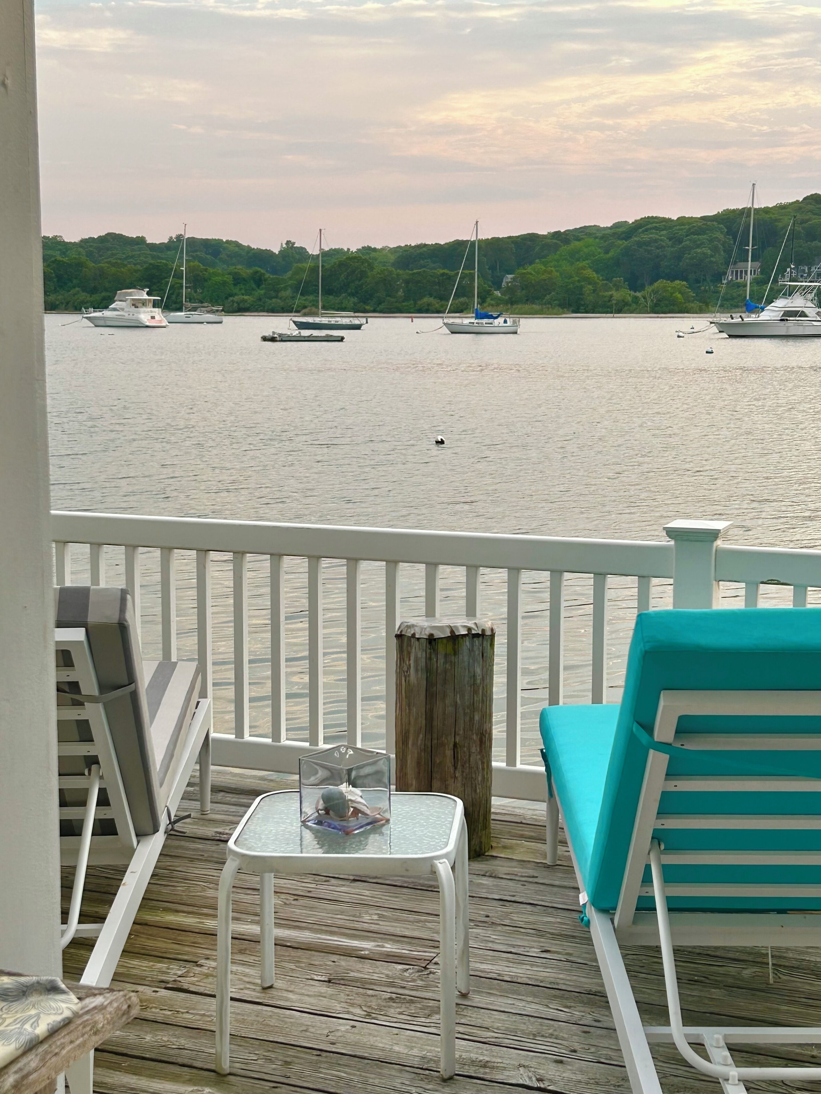 A wooden deck with a glass table and chairs, overlooking a calm body of water with boats.
