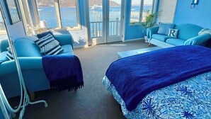 A room with a large bed, a blue sofa, and a desk. There are large windows with a view of the water and buildings. - Waterfront Studio Beach Cottage Wow Sunsets Walk to Village Shops Winery Brewery (Northport)