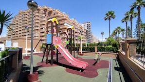 Un parque infantil con un tobogán rojo y columpios, rodeado de palmeras y un edificio grande al fondo.