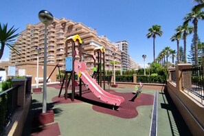 Ein Spielplatz mit einer roten Rutsche und Schaukeln, umgeben von Palmen und einem großen Gebäude im Hintergrund.