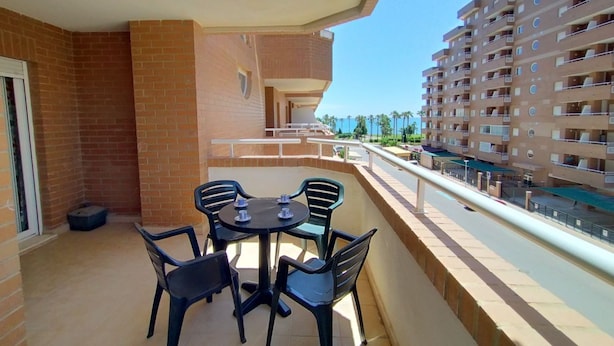 Un balcón con mesa y sillas, con vista a una calle y un edificio de varios pisos.