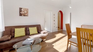 Uma sala de estar com um sofá marrom, uma mesa de centro de vidro, uma mesa de jantar de madeira com cadeiras e uma porta branca com um arco vermelho.