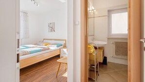 Quarto com cama, mesa de cabeceira e vista para o banheiro, onde se vê uma pia e uma janela.