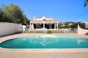 A swimming pool with a white fence, a covered pavilion, and a view of houses in the background.