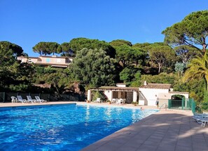 Une piscine avec des chaises longues, un espace salon couvert et une vue sur les arbres et les bâtiments en arrière-plan.