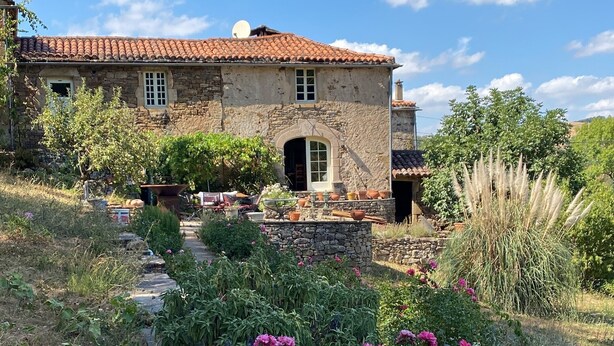 Une maison en pierre avec un toit de tuiles, un chemin en pierre et un jardin où poussent des fleurs roses.