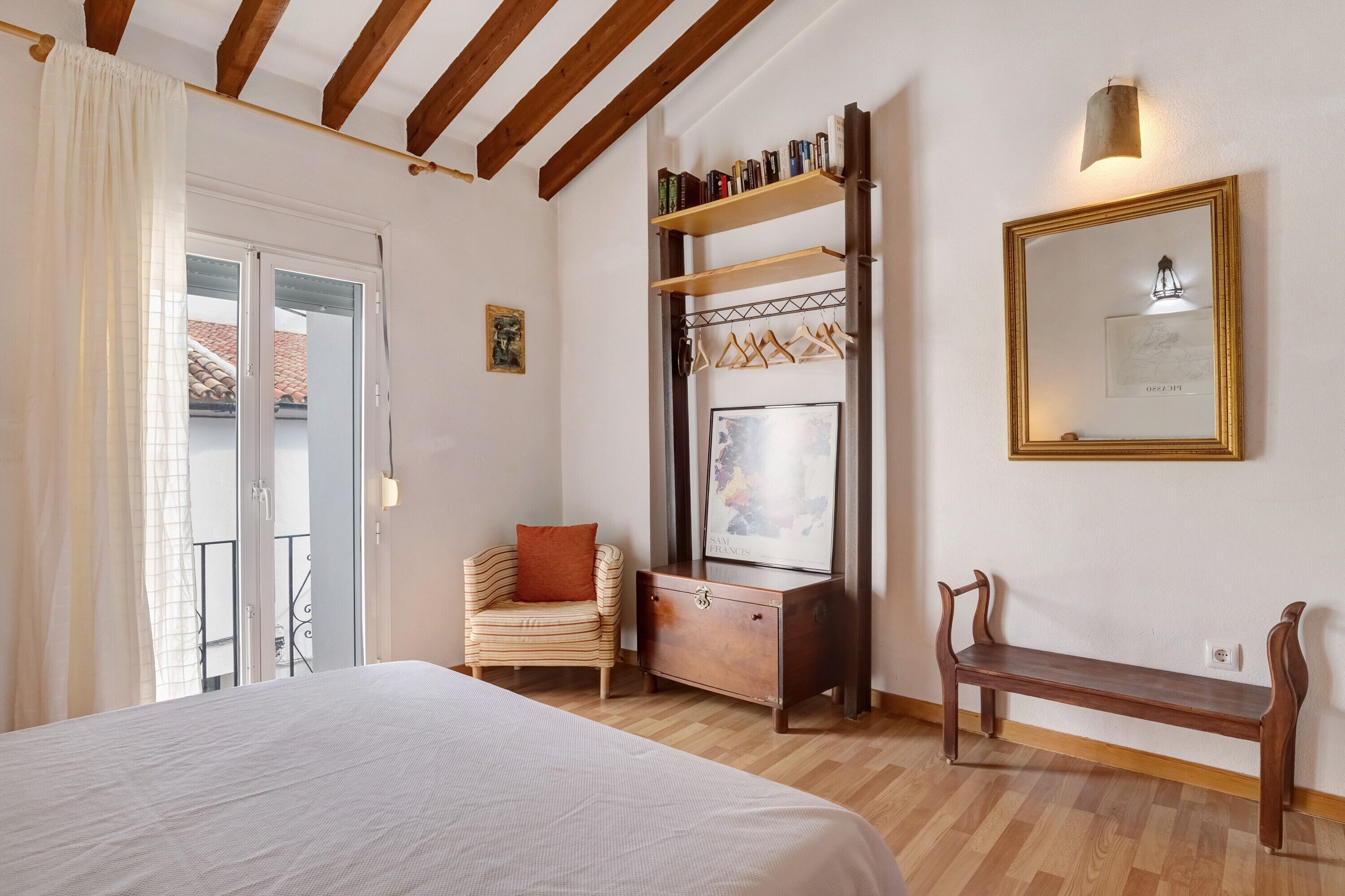 Un dormitorio con cama, una silla, un tocador, un banco y una estantería de pared con libros.