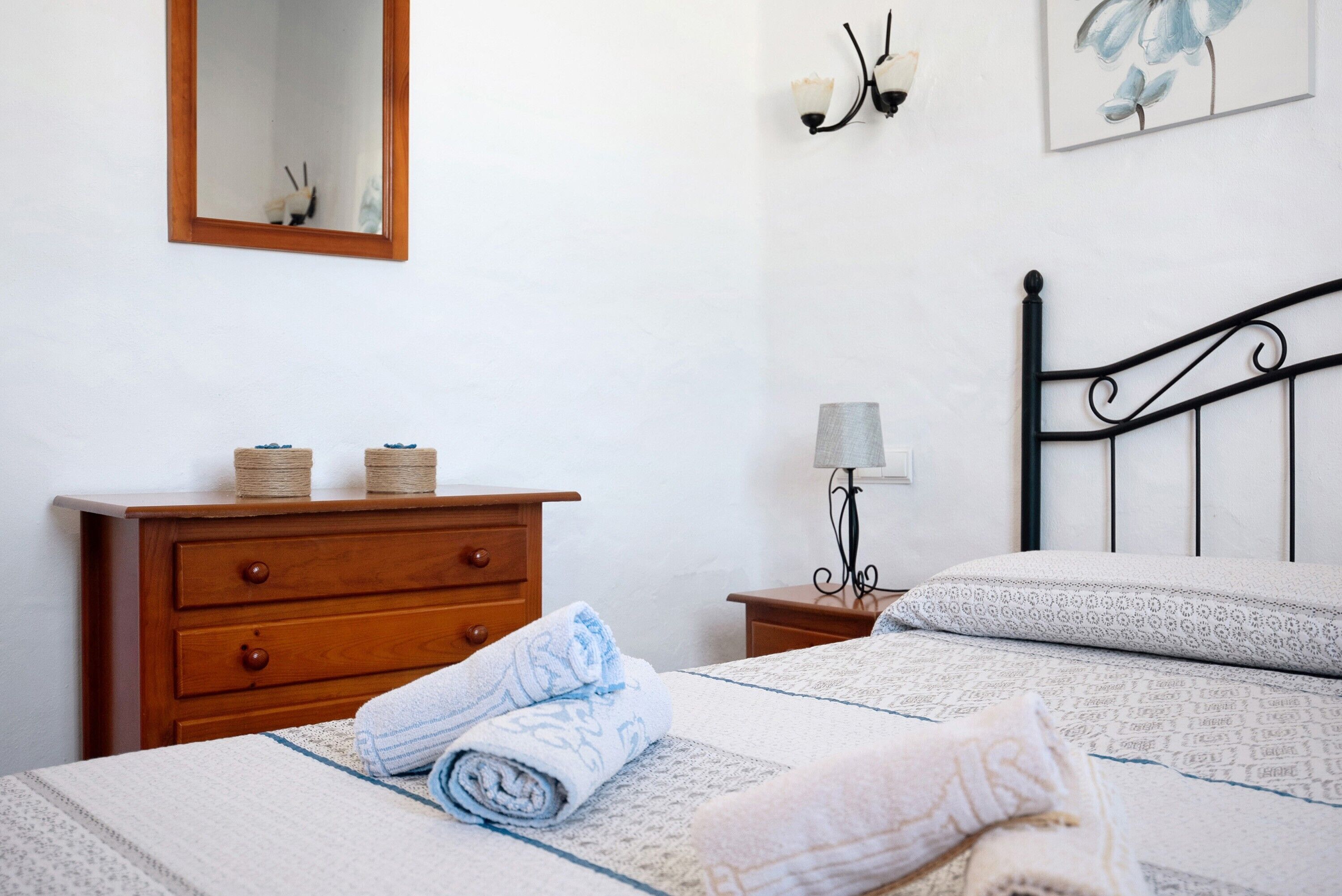 A neatly made bed with a patterned bedspread, a wooden dresser with two spools of thread, a small bedside table with a lamp, and a wall-mounted light fixture.